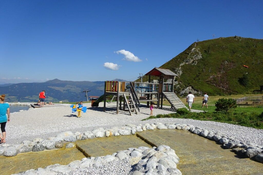 A playground with a slide and climbing structures, surrounded by a rocky area with a clear view of distant mountains.