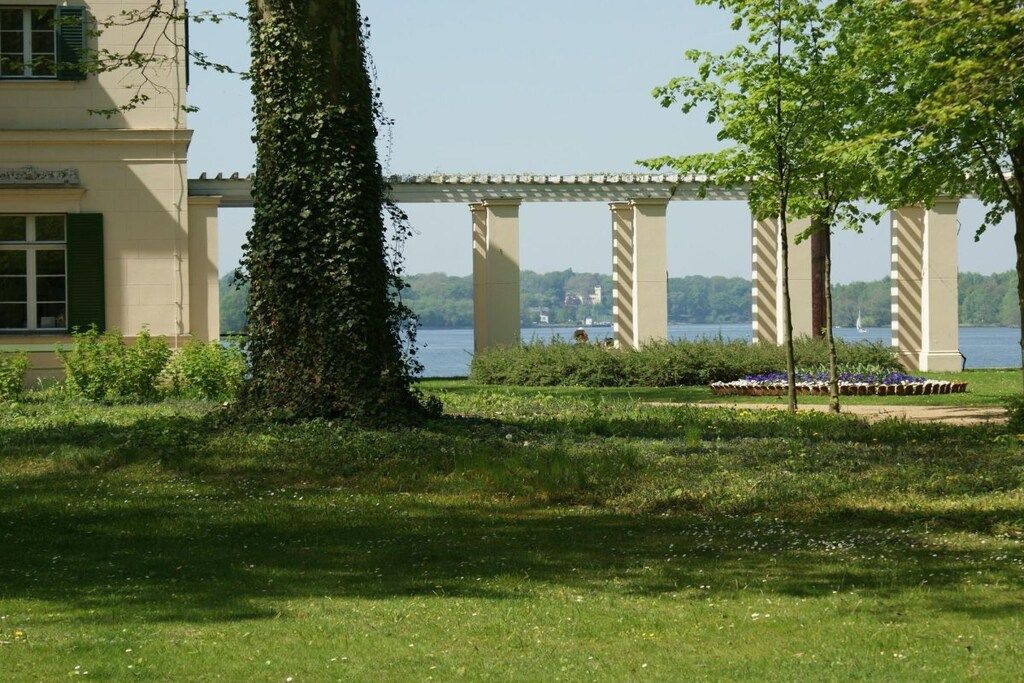 A garden with a pergola, a body of water, and a building with a balcony.