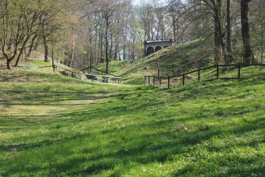 A grassy pathway leading to a structure with arches, surrounded by trees and a fence.
