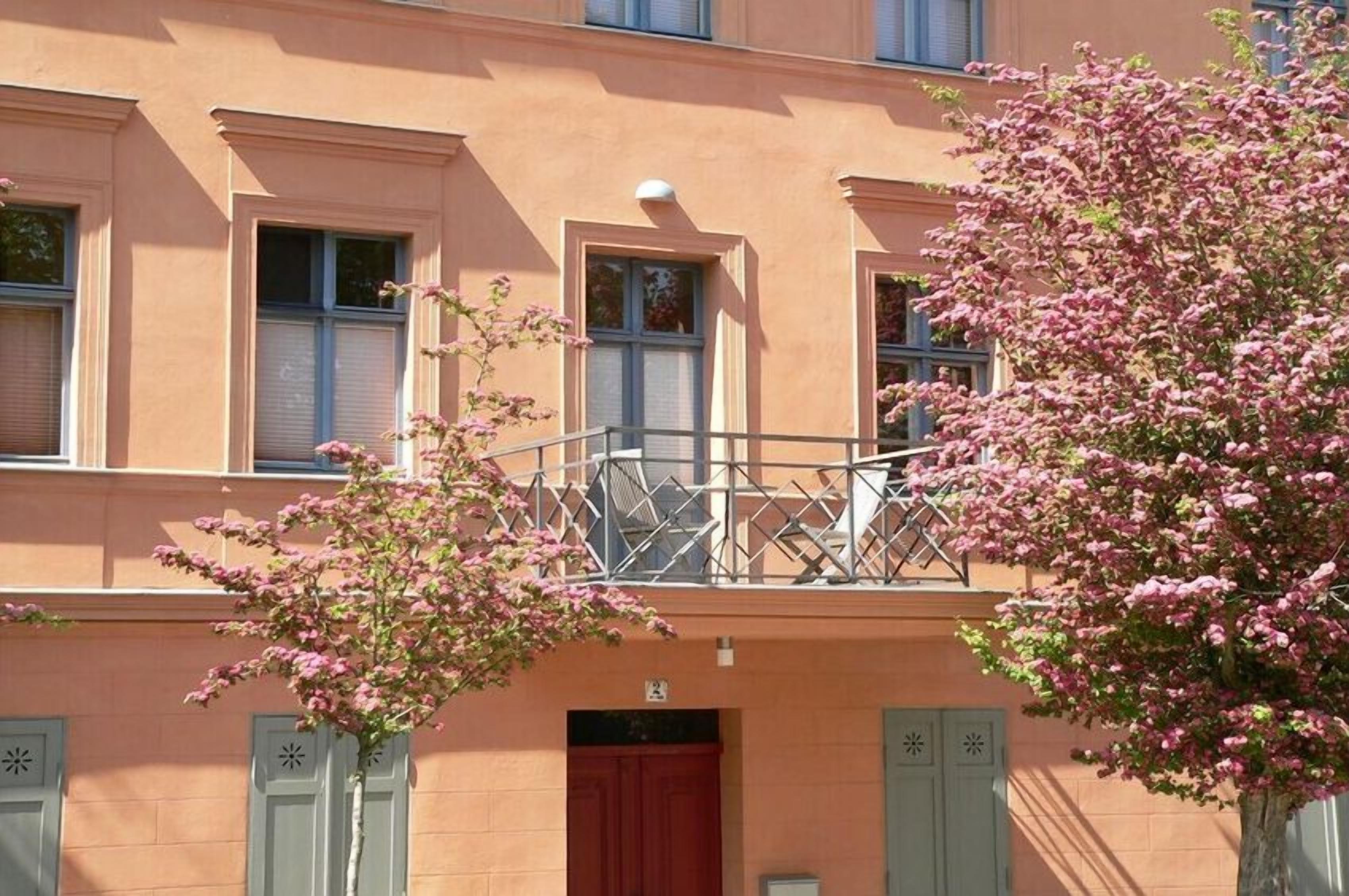 En bygning med balkon, rød dør og et blomstrende træ i forgrunden.