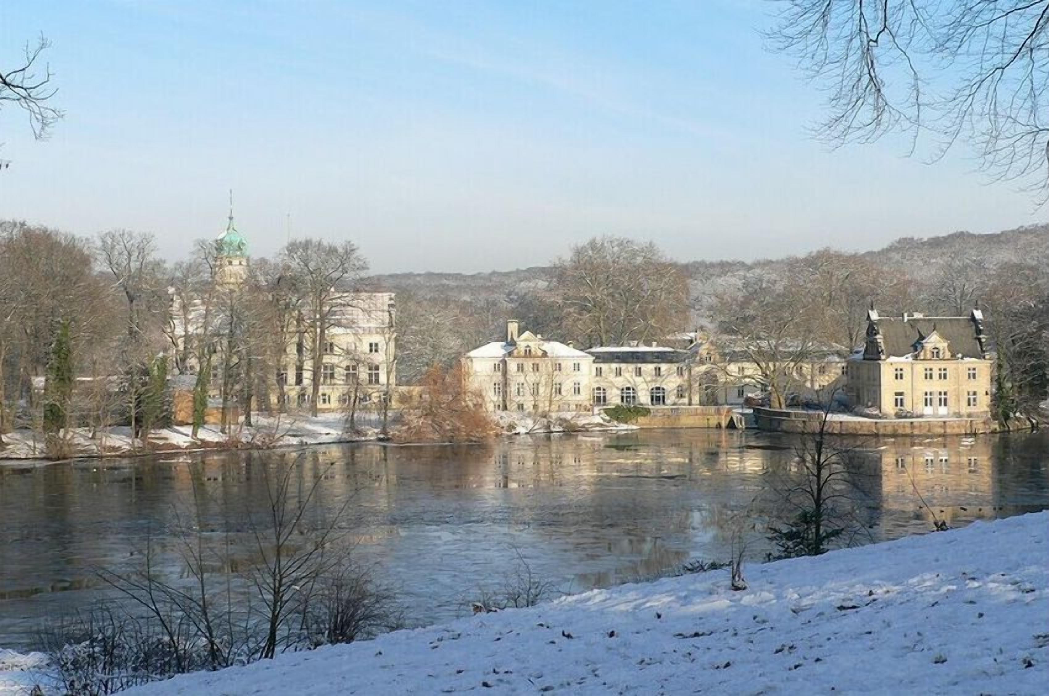 A riverside view of a historic building complex with multiple structures, including a prominent tower with a green roof.