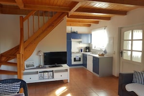 A compact living space with a wooden staircase, a kitchen area with blue cabinets, a dining area with a table and chairs, and a television on a white stand. - Holiday apartment with 1 bedroom (Dierhagen)