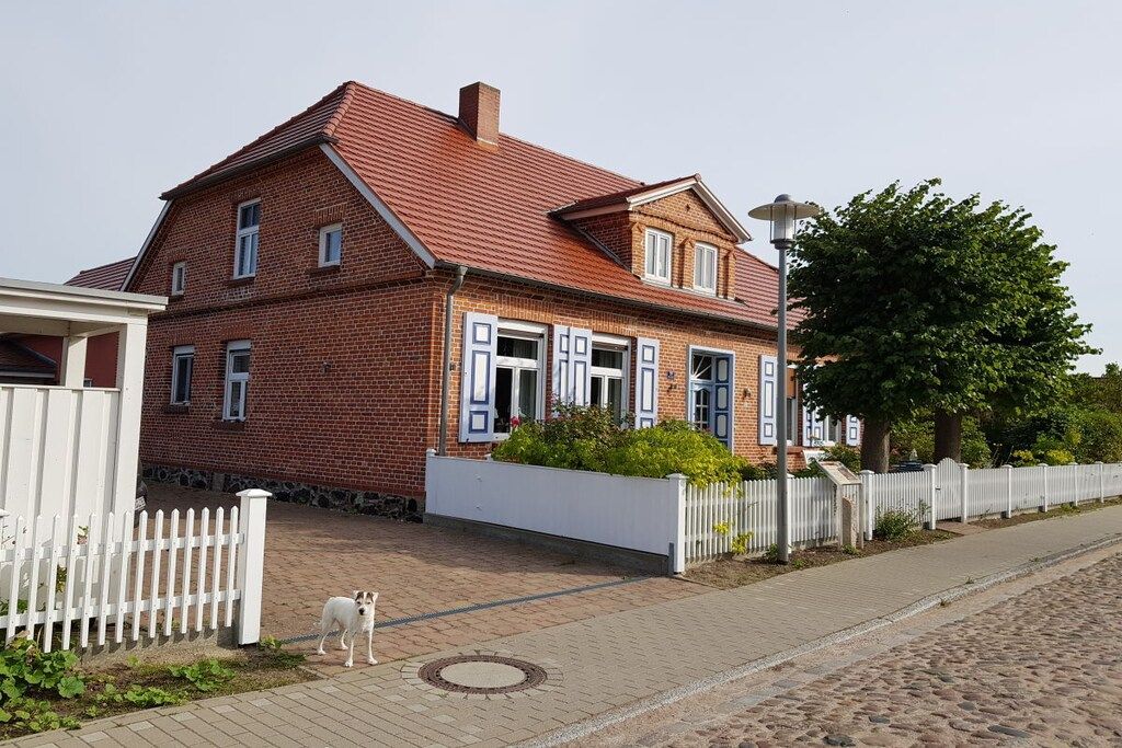 Une maison en briques rouges, avec une clôture en bois peinte en blanc, une allée pavée et un chien qui se tient sur le trottoir.
