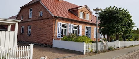 A red brick house with a white picket fence, a paved driveway, and a dog standing on the sidewalk.