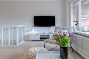 A living room with a flat-screen TV mounted on the wall, a white side table with a lamp, a beige sofa, and a vase with flowers. - Friesenkoje in Mellhörn (List)