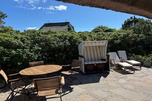 An outdoor patio with a round wooden table, cushioned chairs, and a covered seating area. - Friesenkoje in Mellhörn (List)