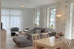 A modern living room with a sofa, ottoman, dining table, and chairs. There are curtains, a lamp, and a window with a view. - No. 9 in Villa Strandburg (Ostseebad Binz)