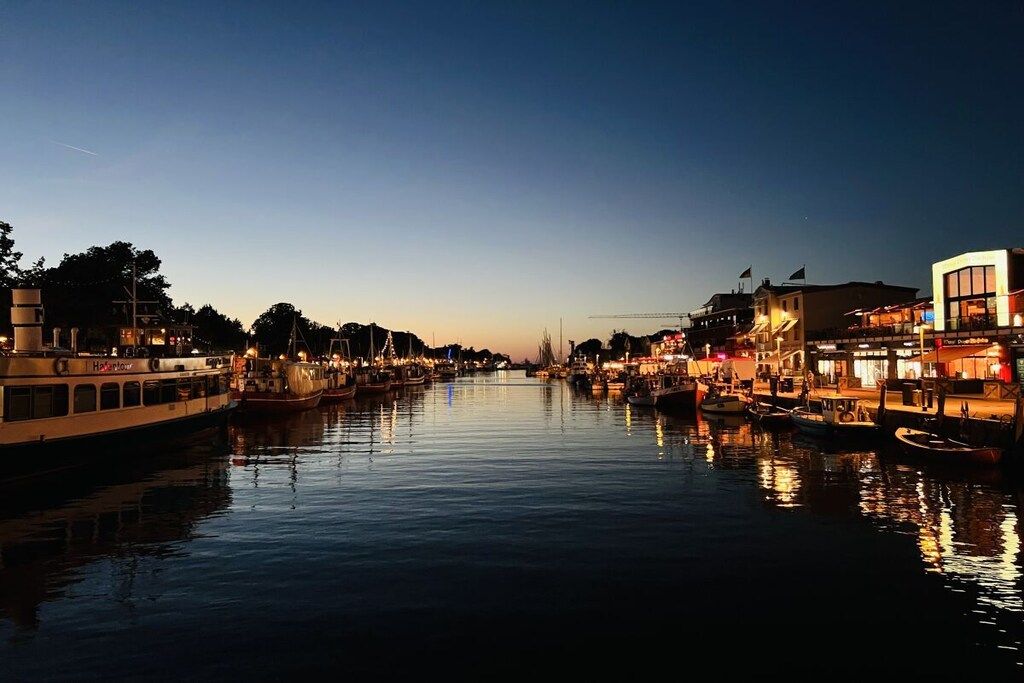 A waterfront cityscape with boats docked along the river, buildings with illuminated signs, and a clear sky.