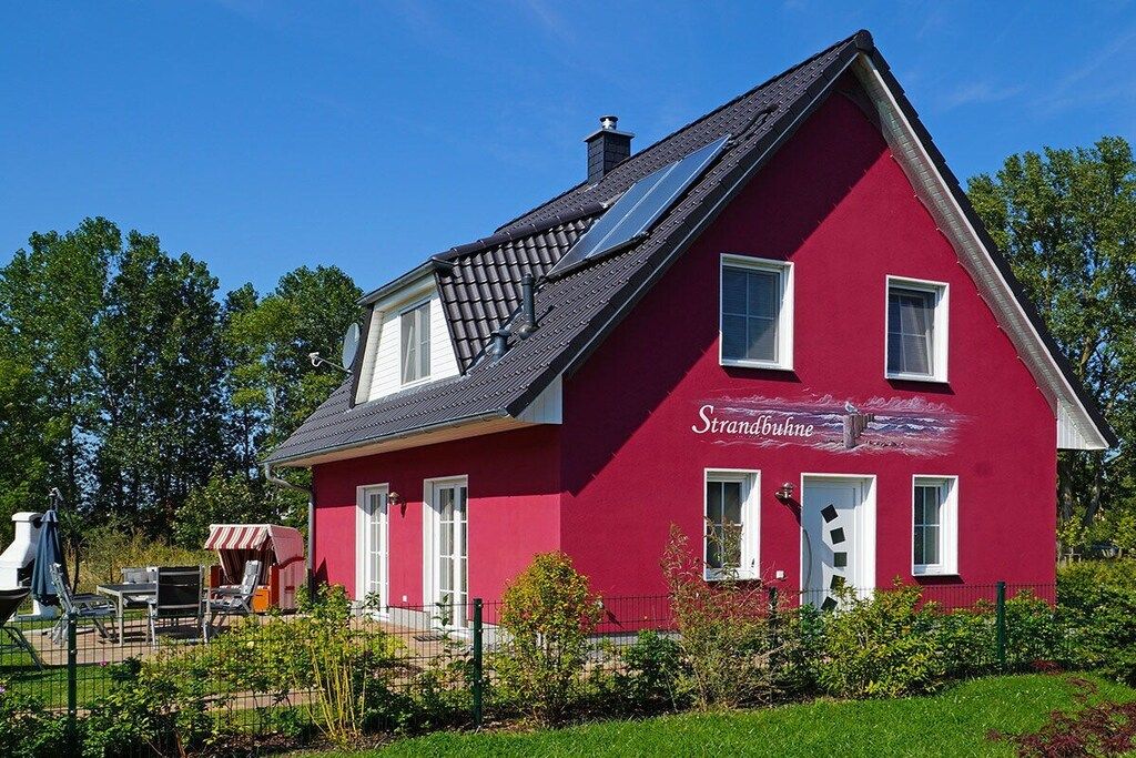 A red house with a sloped roof, solar panels, and a fenced yard with outdoor furniture.