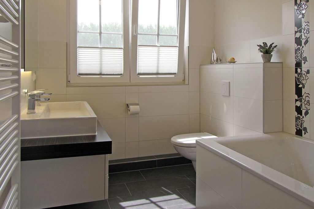 A bathroom with a toilet, a sink, and a bathtub. There is a window with blinds, a towel rack, and a potted plant on the counter.