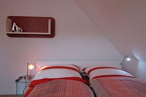 A bedroom with two beds, a bedside table with a lamp, and a shelf with decorative items. - Holiday home with 3 bedrooms (Glowe\/Rügen)
