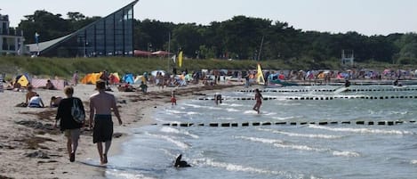 A beach with people walking and swimming, tents, and a modern building in the background.