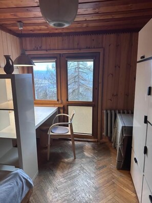 A cabin interior with a wooden ceiling, a dining table with a chair, a window with a view of trees, and a radiator.