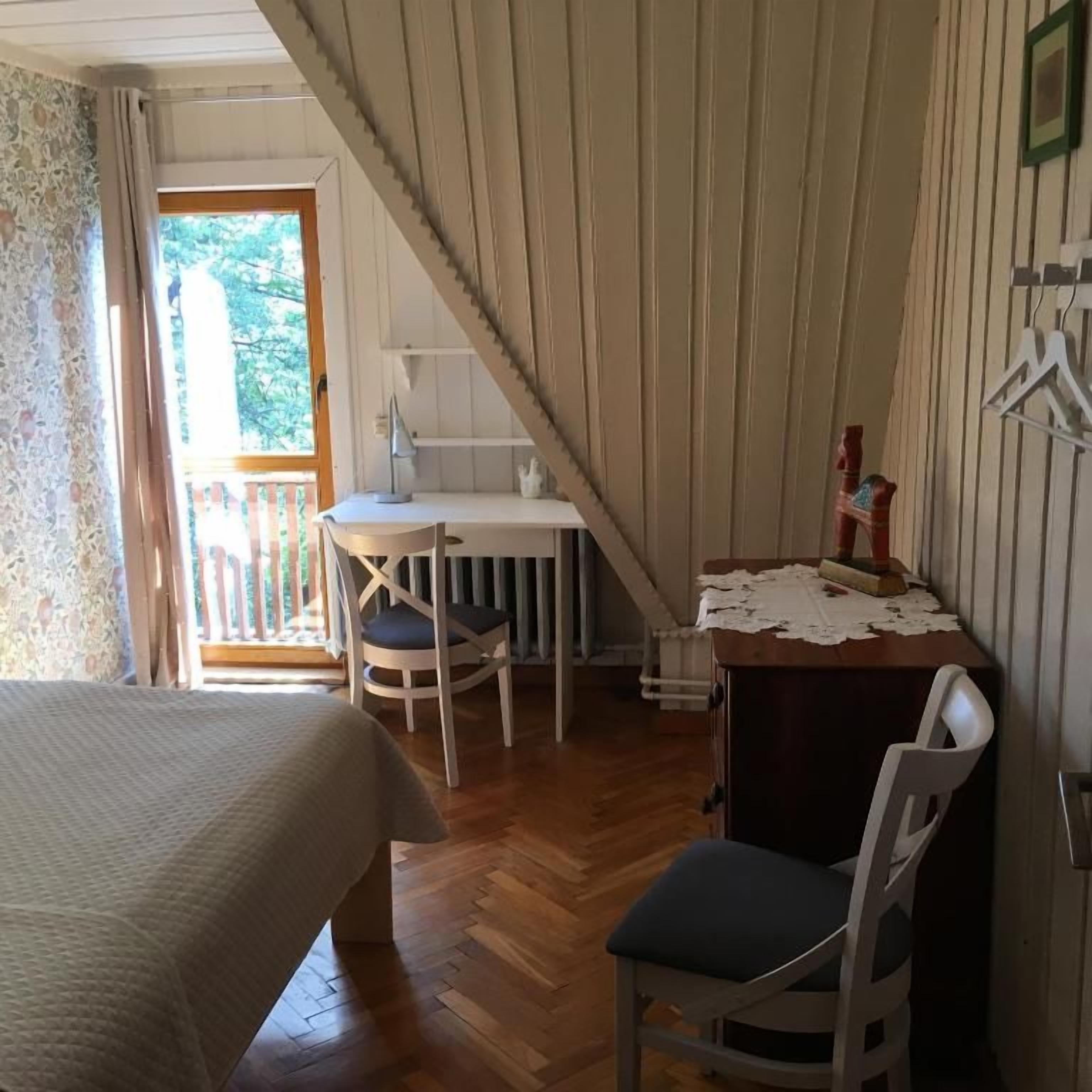 A bedroom with a bed, a desk, and a chair. There is a window with curtains, a door leading to a balcony, and a coat rack.
