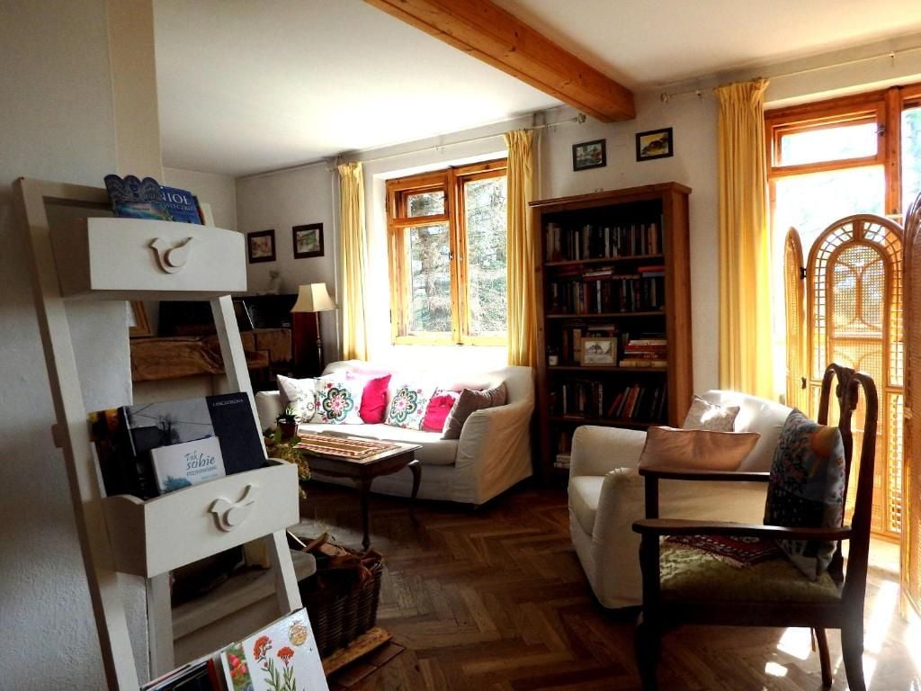 A cozy living room with a white sofa, a wooden chair, a bookshelf, and a window with curtains.