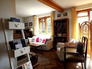 A cozy living room with a white sofa, a wooden chair, a bookshelf, and a window with curtains.