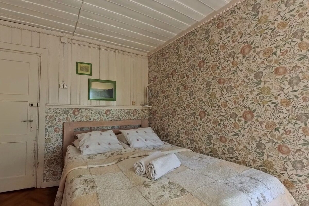 A bedroom with a wooden door, a bed with a floral-patterned headboard, and a floral wallpaper on one wall.