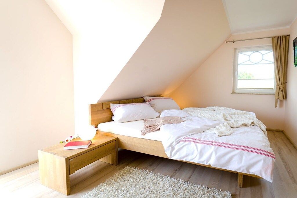 Une chambre avec un plafond en pente, un lit avec une literie blanche, une table de chevet et une fenêtre avec des rideaux.