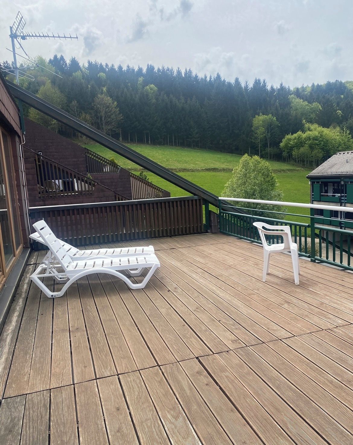 Eine Holzterrasse mit zwei weißen Liegestühlen und Blick auf ein grünes Feld und Bäume.