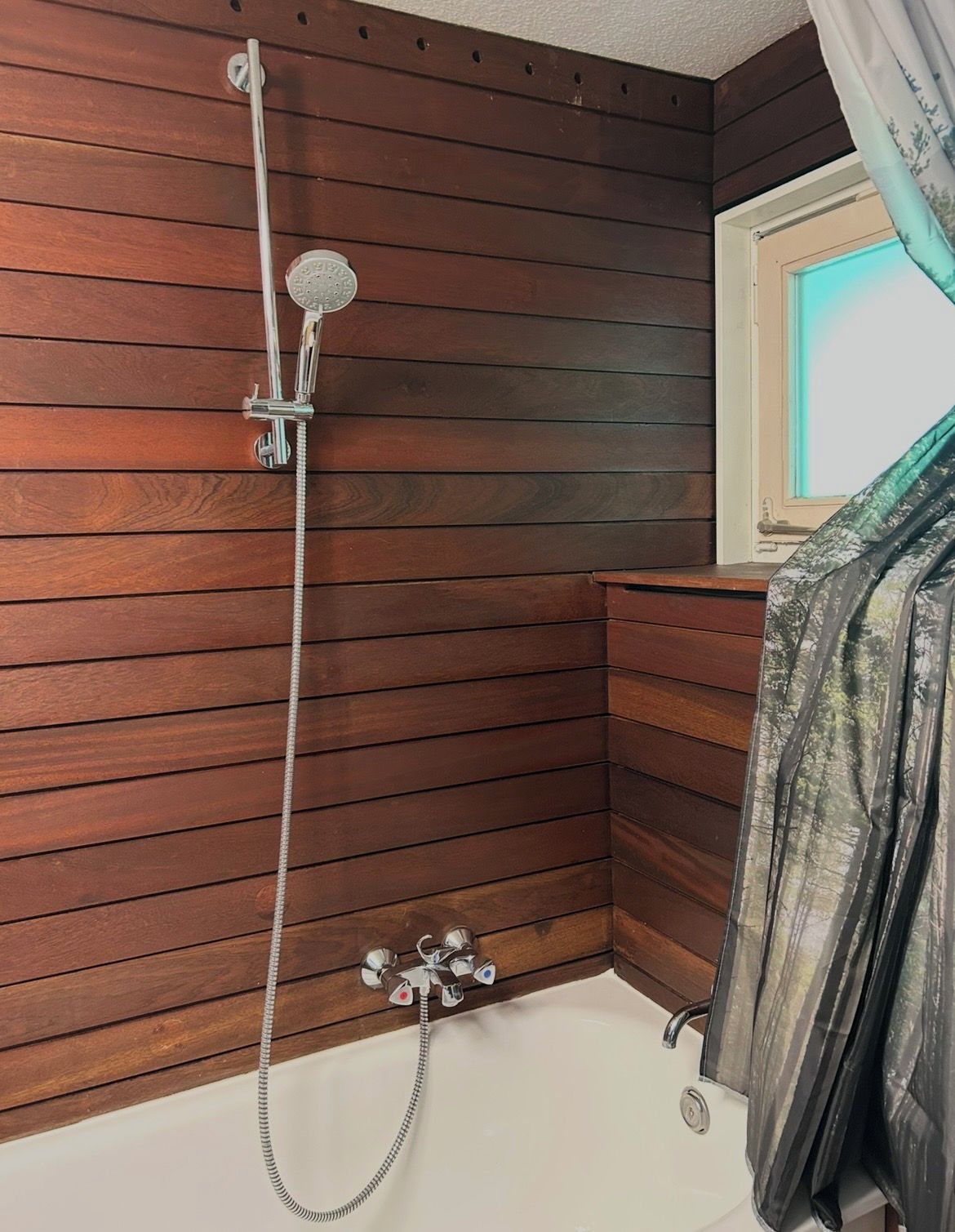 Ein Badezimmer mit Holzvertäfelung, einer Dusche inklusive Duschbrausehandstück und einem Fenster mit Vorhang.