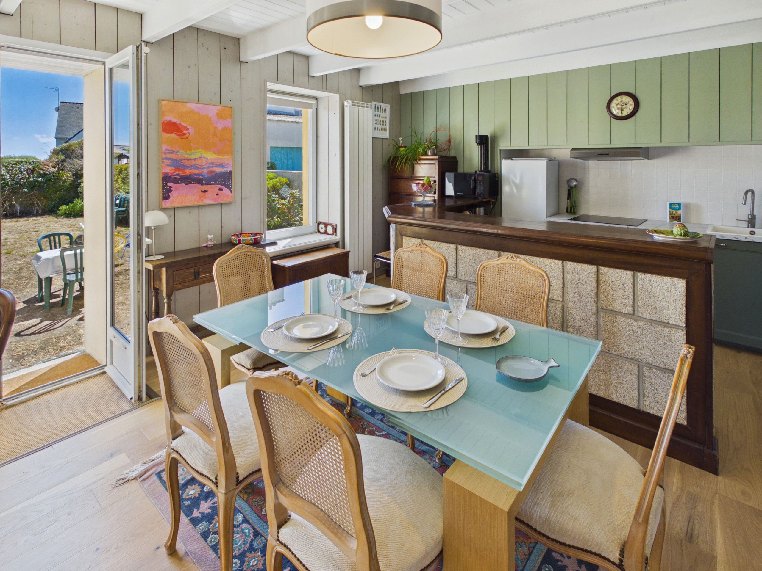 A dining area with a table set for four, surrounded by wicker chairs, a kitchen with a green cabinet, and a view through an open door.