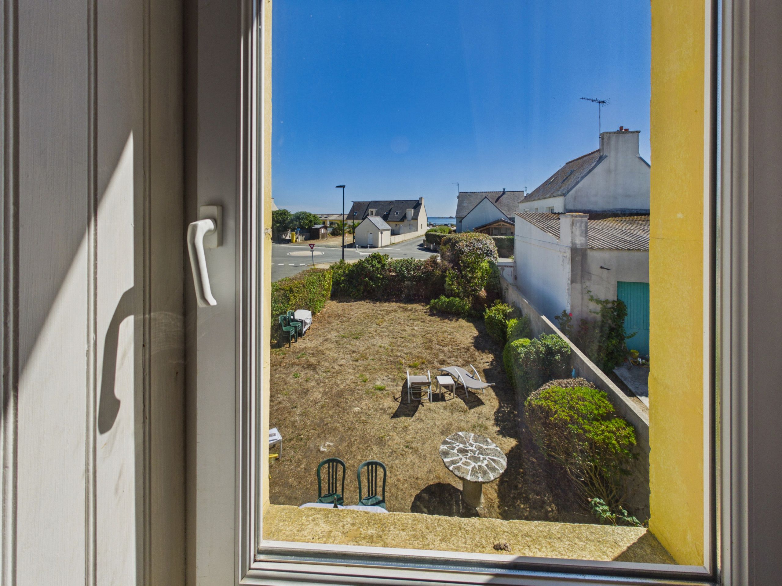Une vue à travers une fenêtre donnant sur une cour intérieure avec une table et des chaises, un petit jardin et des bâtiments en arrière-plan.