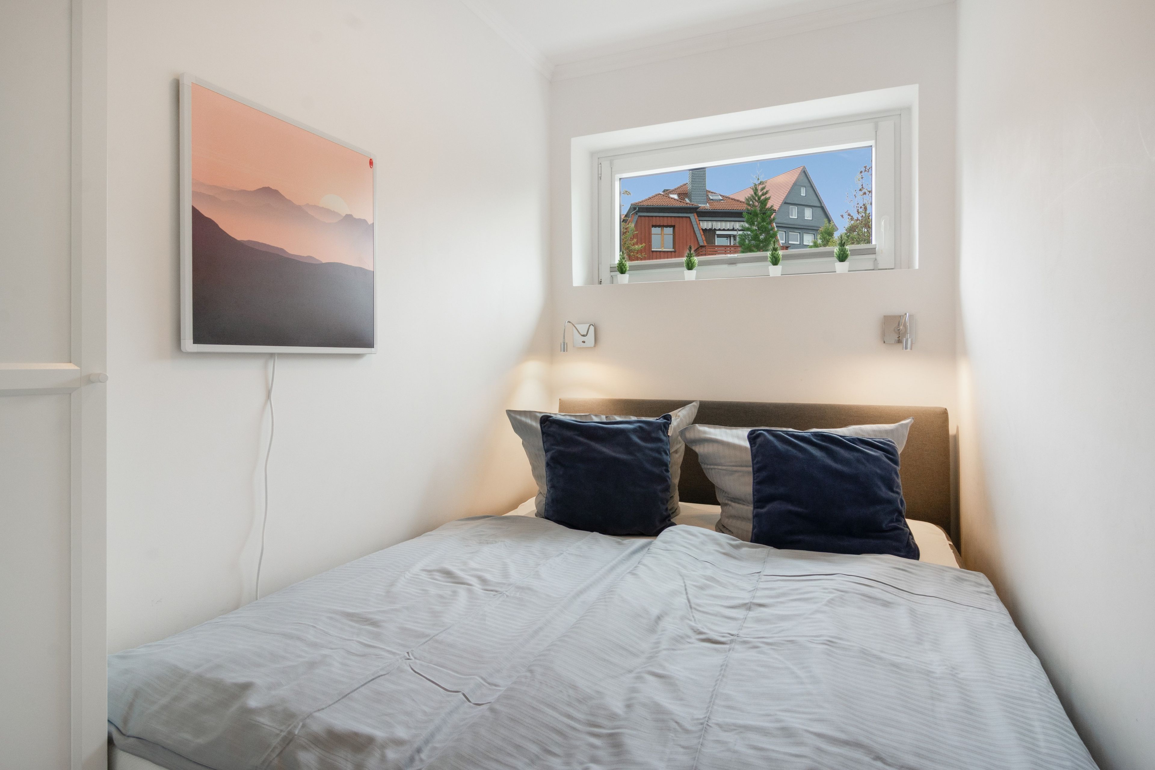 Et soveværelse med seng, puder, et vindue med udsigt til et hus, og et billede hængt på væggen.