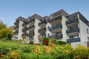 Ein modernes, mehrstöckiges Wohngebäude mit Balkonen, umgeben von einem begrünten Garten mit verschiedenen Sträuchern und Bäumen.