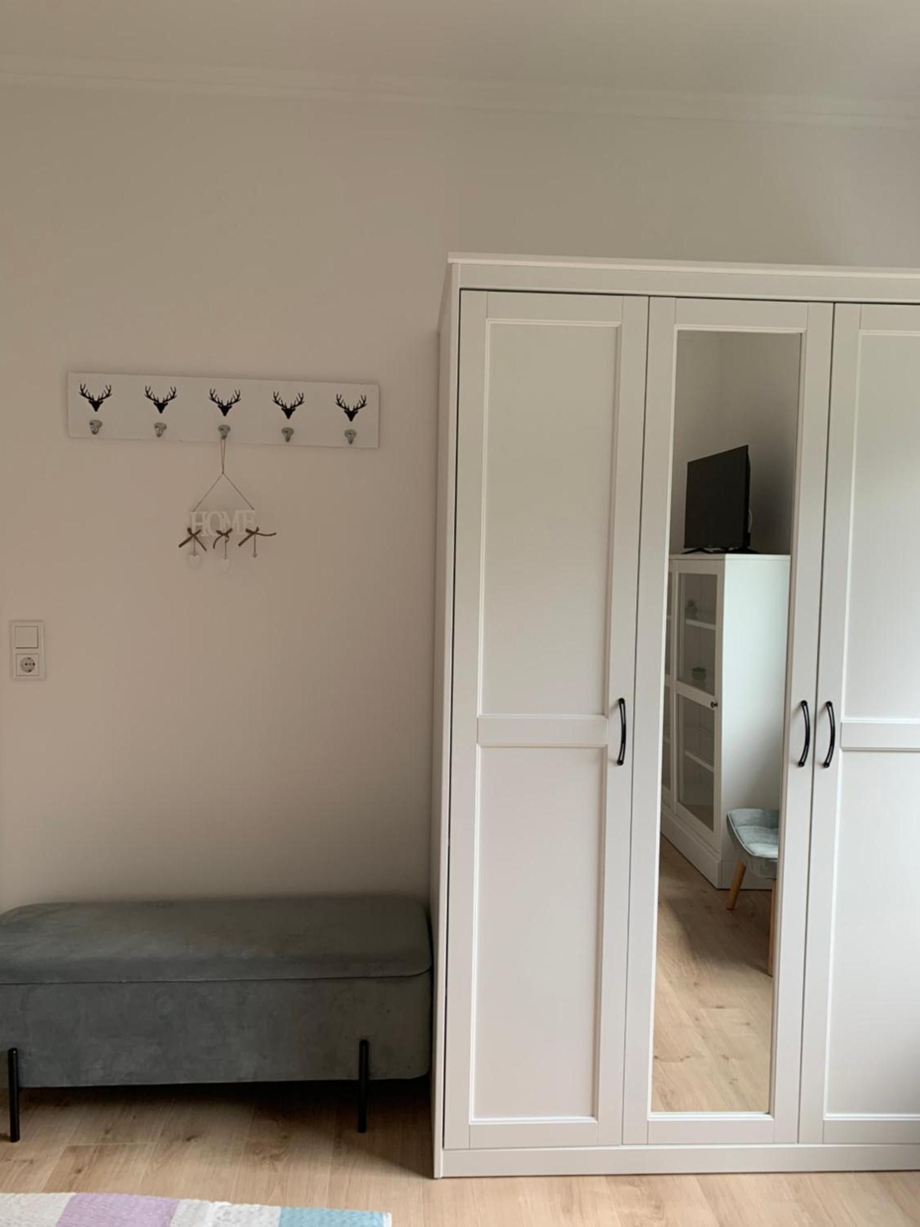 Une pièce avec un banc gris, une armoire blanche et un porte-manteau orné de bois de cerf et d’une pancarte indiquant le mot « MAISON ».