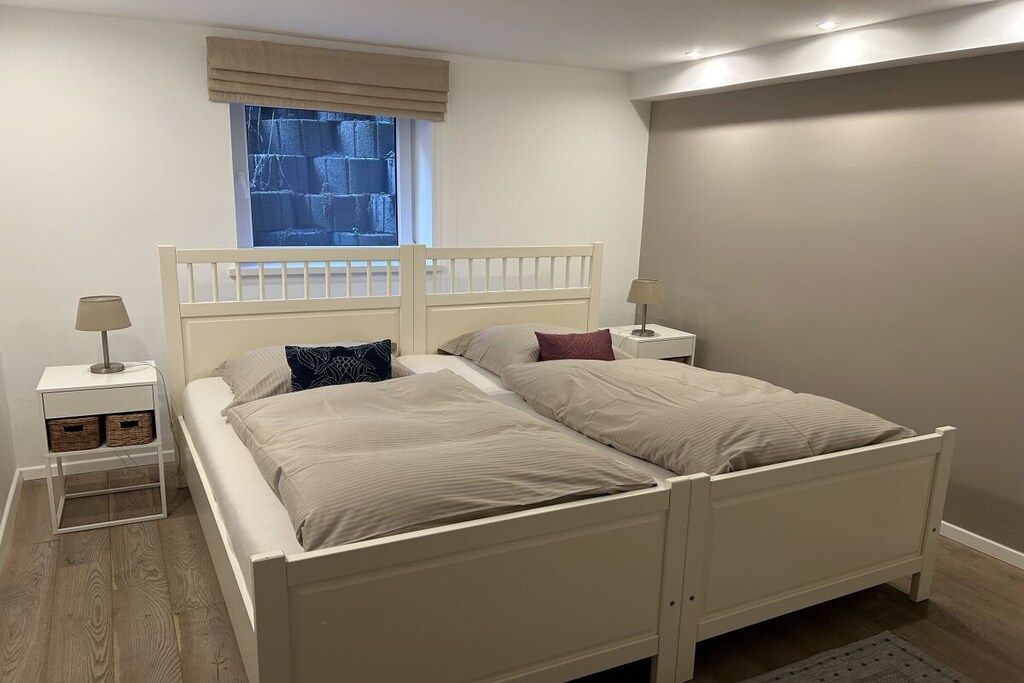 A double bed with a headboard, two bedside tables, a window with blinds, and a plain wall.