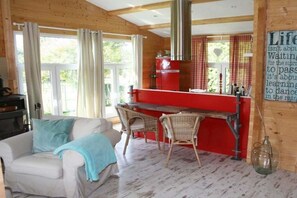 A cozy room with a white sofa, two wicker chairs, a red kitchen area, and a large window with curtains. - Holiday home Rainbow (Friedrichskoog)