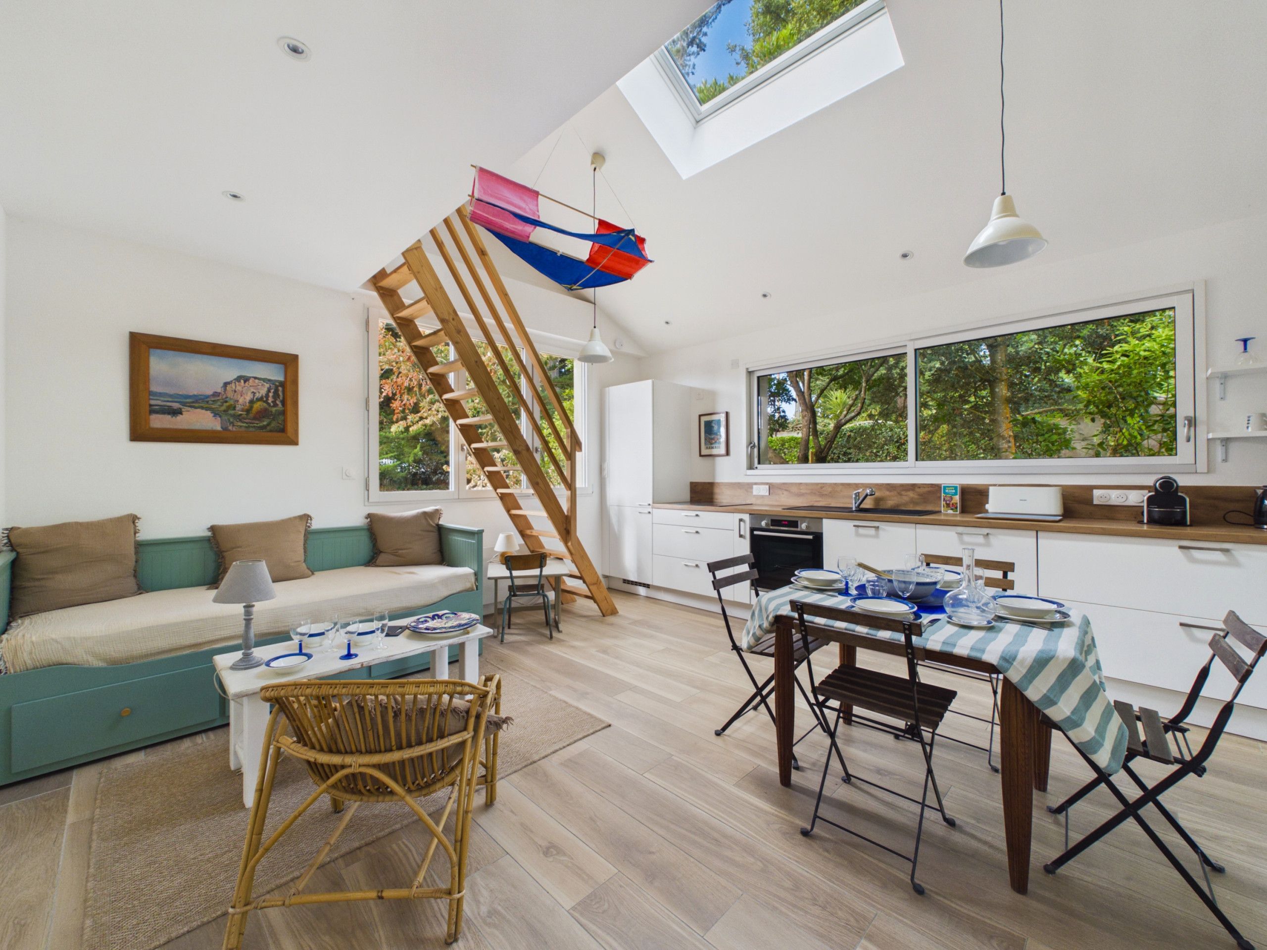 A modern kitchen with a wooden staircase, a dining area with a table set for two, and a large window overlooking greenery.