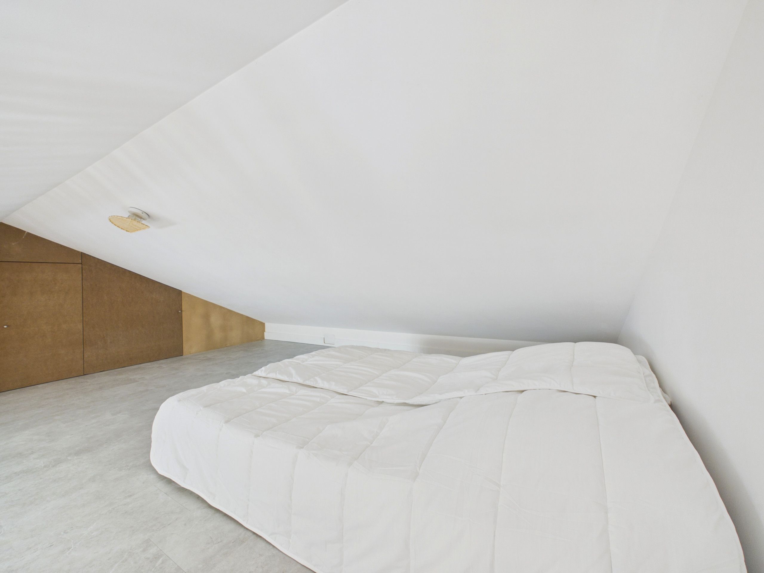 Ein Schlafzimmer mit schrägem Dach, einem Bett mit weißer Bettdecke und einer Holzwand links.