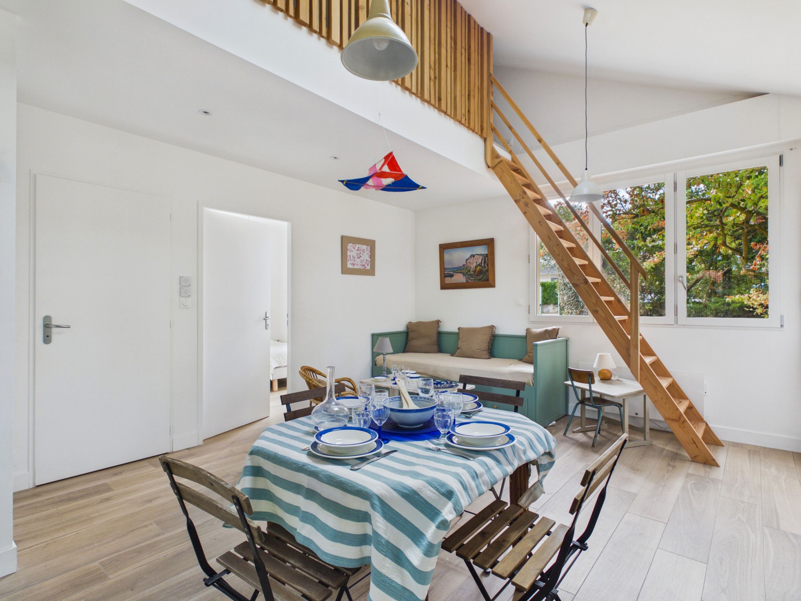 Un comedor con una mesa lista para cuatro, una escalera de madera y vistas a árboles a través de una ventana.