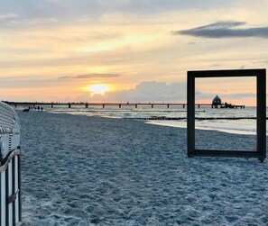 A beach with a pier extending into the water, a sunset in the background, and a framed picture on the sand. - Sünnenkringel Apt. 5 (Zingst)