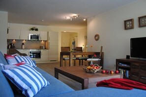 A living room with a sofa, a coffee table with fruit, a TV, and a kitchen in the background. - Holiday apartment with 2 bedrooms (Breege\/ Juliusruh OT Juliusruh)