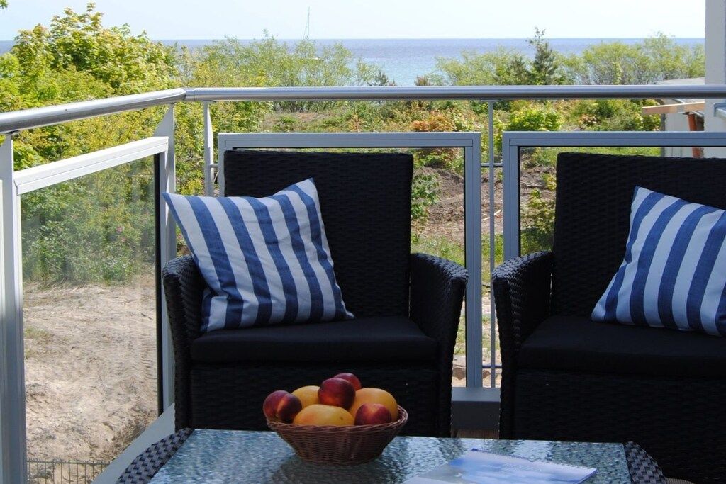 Dos sillas de mimbre con cojines a rayas, una cesta de mimbre con fruta y una mesa en un balcón con vistas al mar.