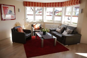 A living room with a sofa, armchair, and coffee table, large windows with curtains, and a radiator. - Holiday apartment with 1 bedroom (Zingst)