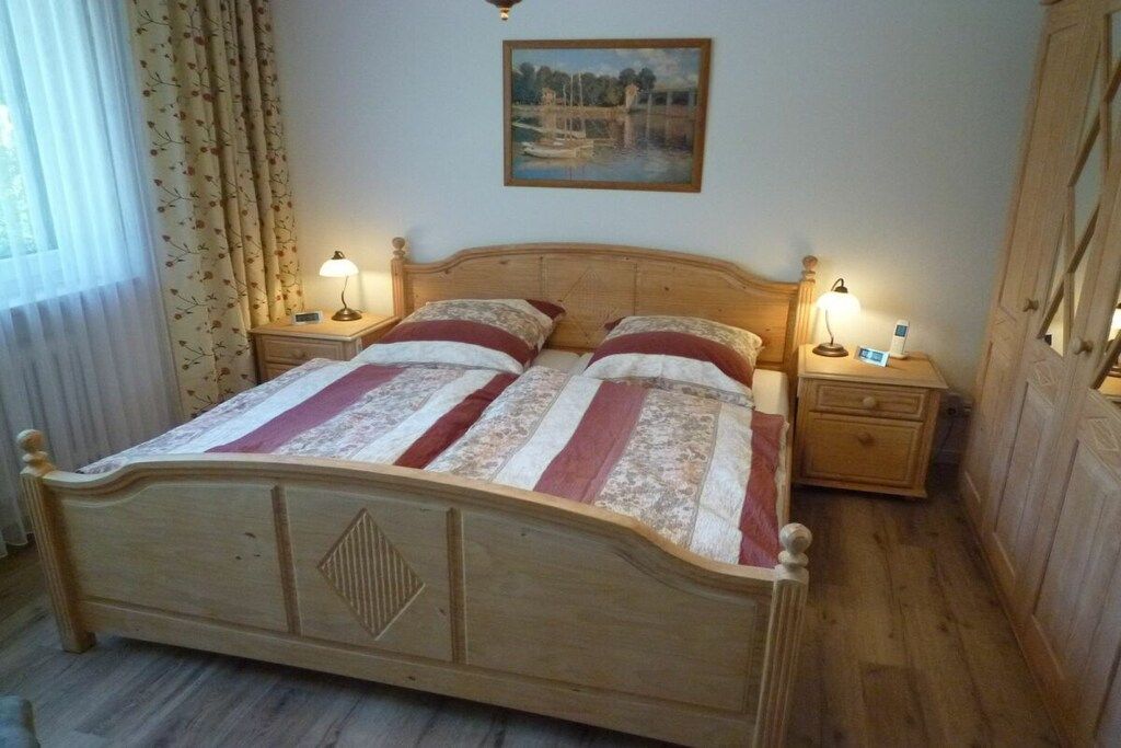 A wooden bed with a headboard, two bedside tables with lamps, a painting on the wall, and a window with curtains.