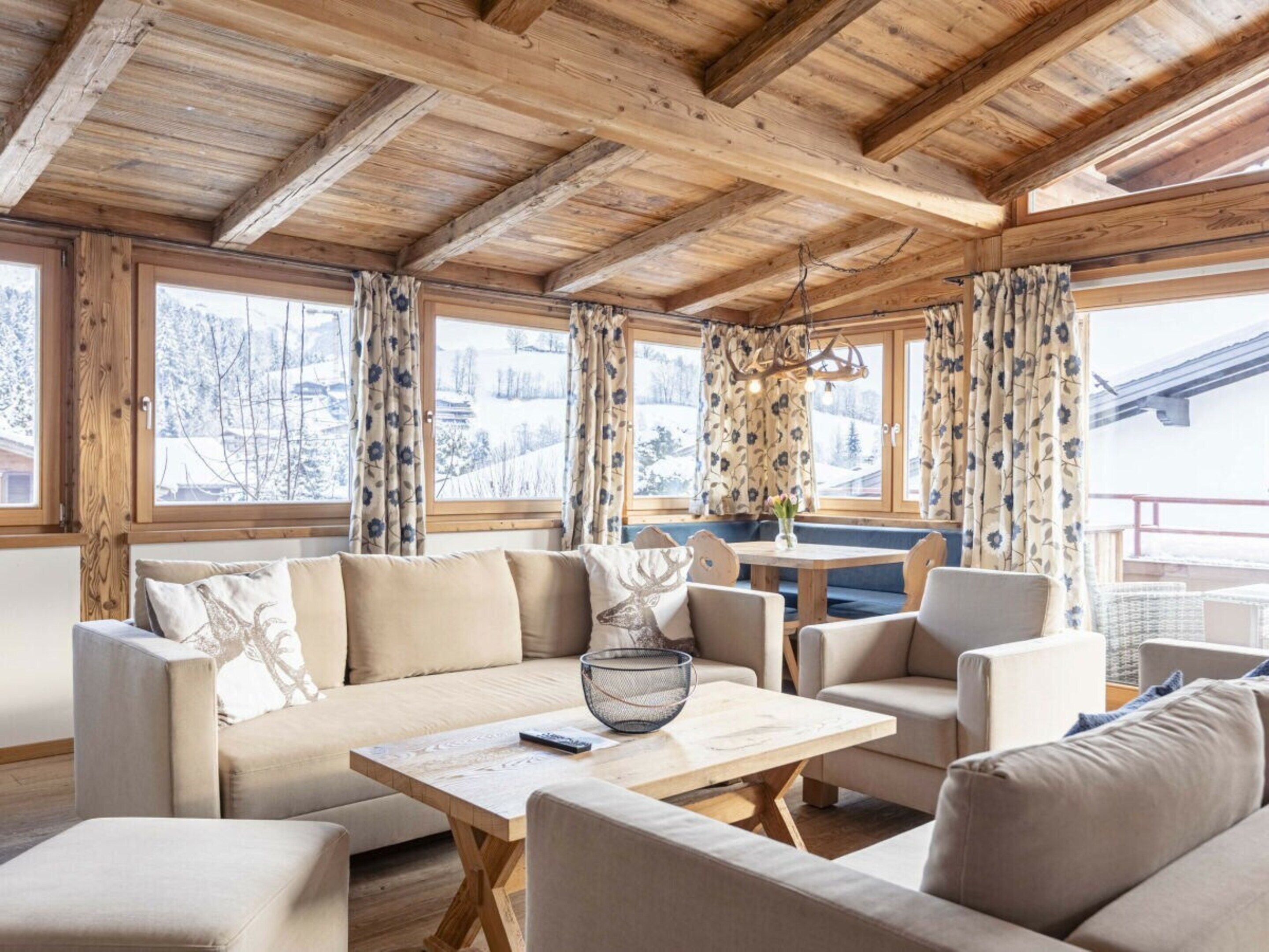 A cozy room with a wooden ceiling, large windows with curtains, a sofa set, and a dining table.