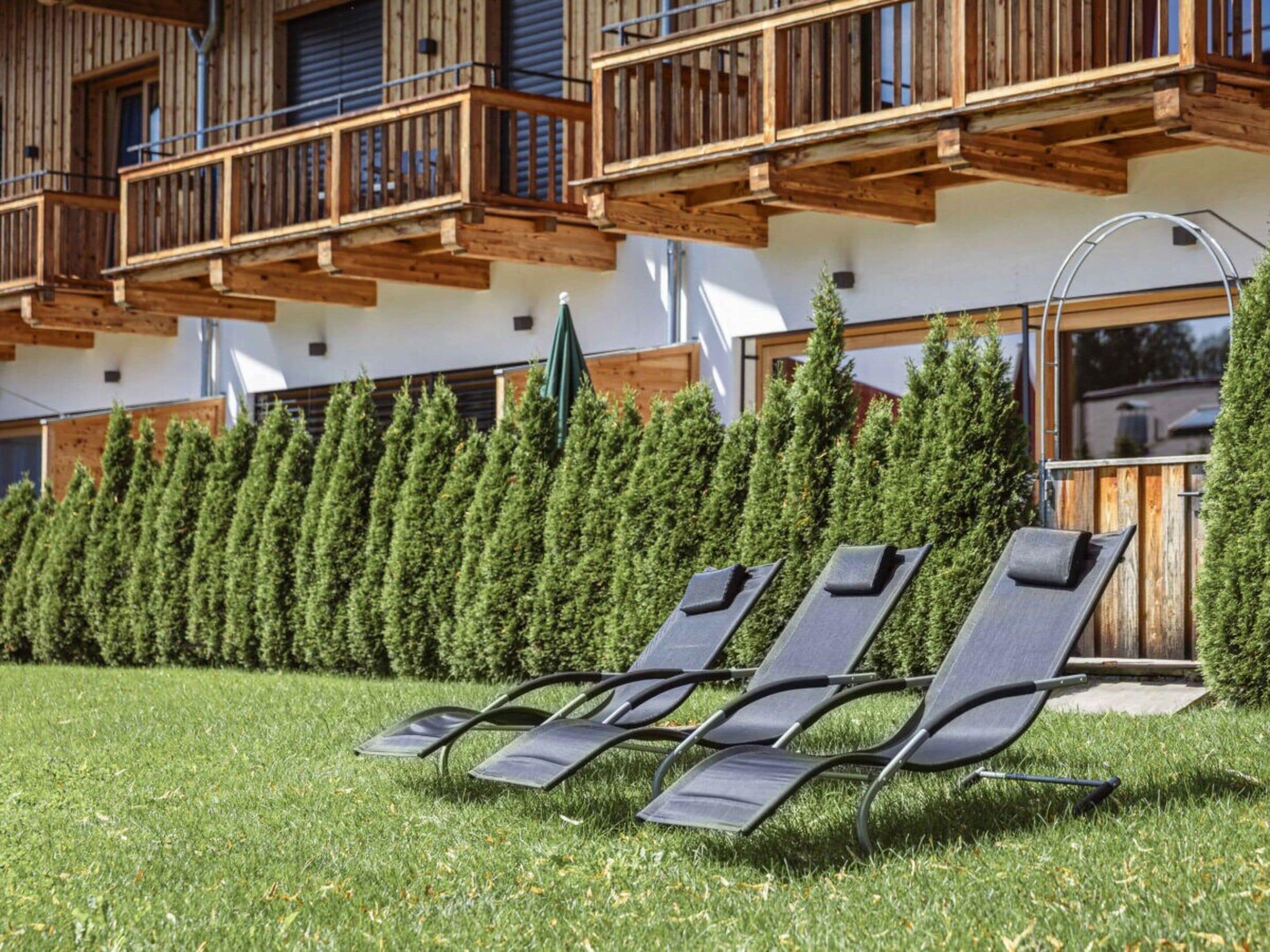 A building with wooden balconies, a row of green coniferous trees, and three sun loungers on a grassy lawn.