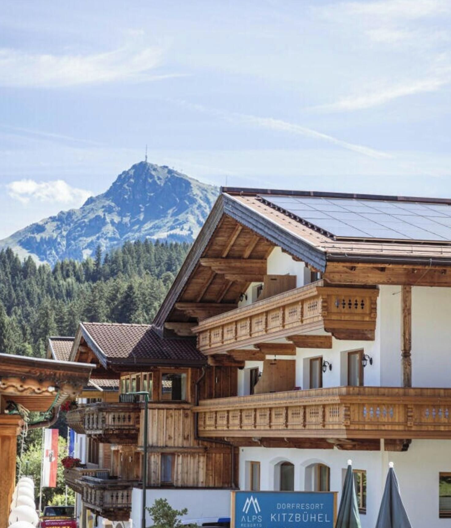 A traditional Alpine chalet with a wooden balcony, a mountain backdrop, and a sign for 'Dorfresort Alps Kitzbühel'.