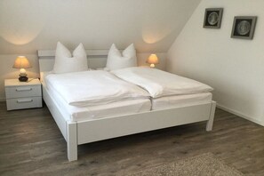 A bedroom with a white bed, two bedside tables with lamps, a wooden floor, and a small rug. - Holiday apartment with 2 bedrooms (Norddeich)