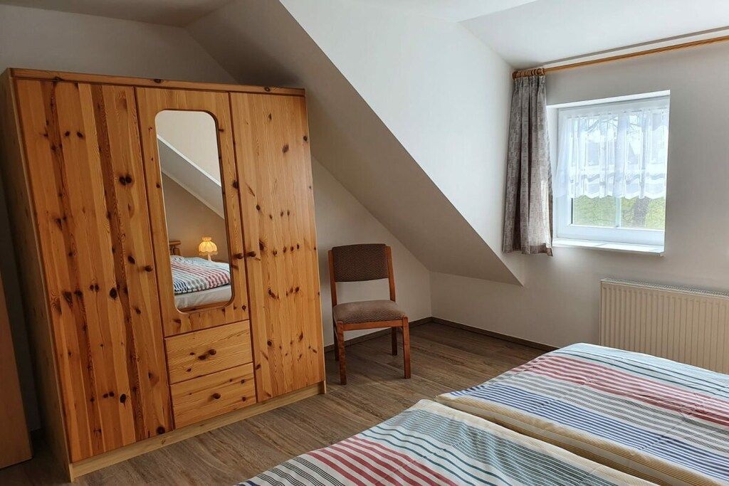 A bedroom with a sloped ceiling, a wooden wardrobe with a mirror, a single bed, a chair, and a window with curtains.