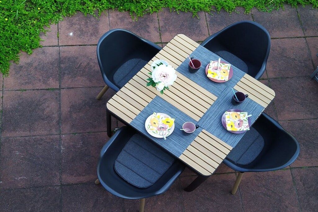 Une table carrée, entourée de quatre chaises, chacune agrémentée d’un coussin. La table est décorée d’un centre de table floral et porte deux assiettes avec des couverts.