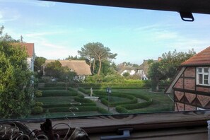 A view from inside a vehicle looking out at a residential area with neatly trimmed hedges, a statue, and traditional houses with thatched roofs. - Holiday apartment Hehl (Born)