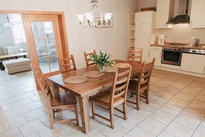 Uma sala de jantar com mesa e cadeiras de madeira, um vaso com flores e uma cozinha ao fundo.