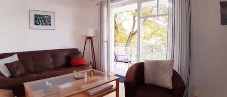 Un salon avec un canapé marron, une table basse en verre et un fauteuil marron. Il y a une fenêtre avec des rideaux, un tableau au mur et une lampe sur pied.