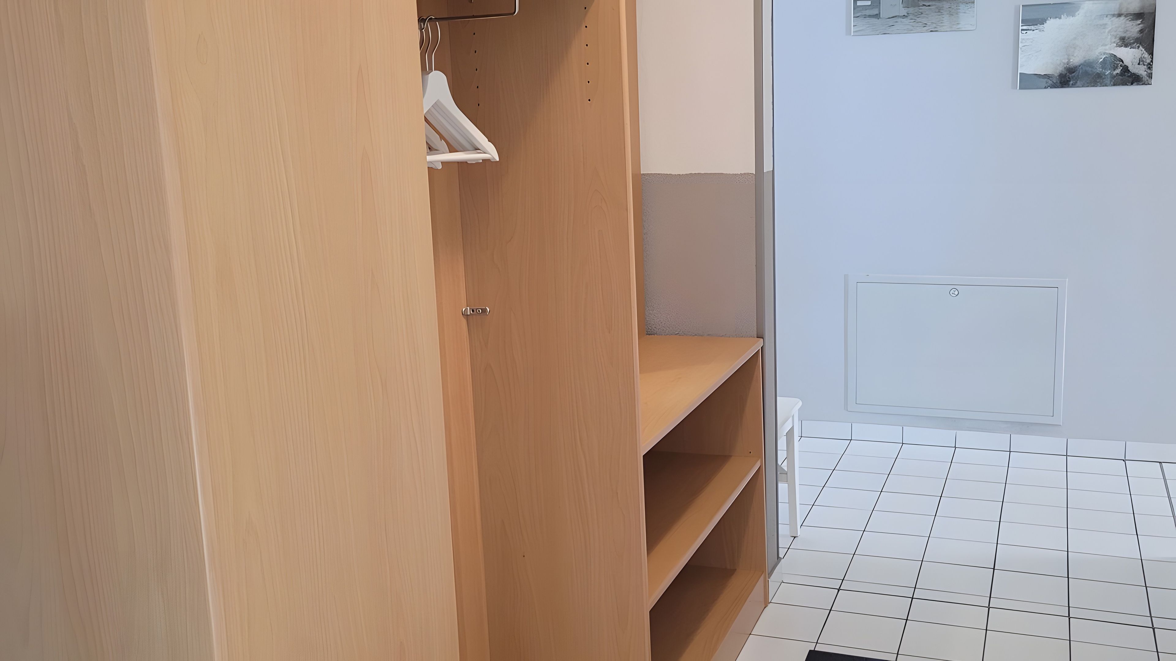 Ein Holzkleiderschrank mit Kleiderstange und Ablage, ein weiß gefliester Boden und ein an der Wand montierter Lichtschalter.