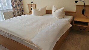 Ein ordentlich bezogenes Bett mit weißen Leinen und Kissen, ein Nachttisch mit Lampe und eine Steckdose an der Wand.